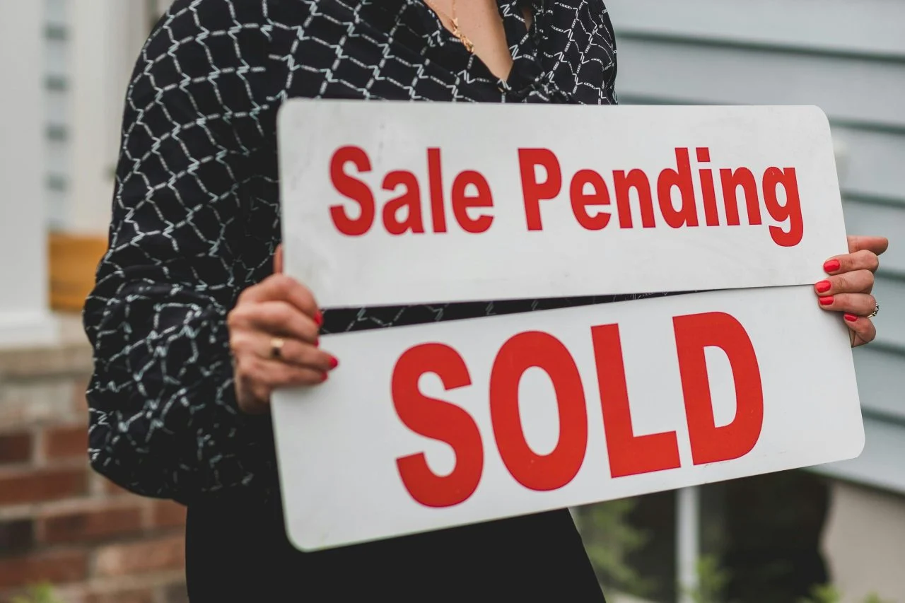 Person holding "Sale Pending" and "SOLD" signs in front of a house, indicating a property has been sold.