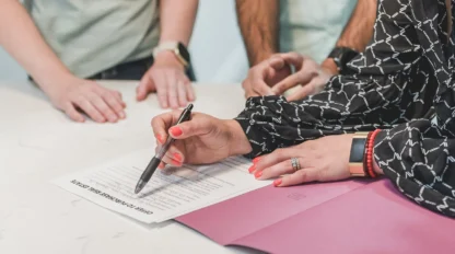 A person is signing a document on a table while three others observe, with a folder and pen visible.