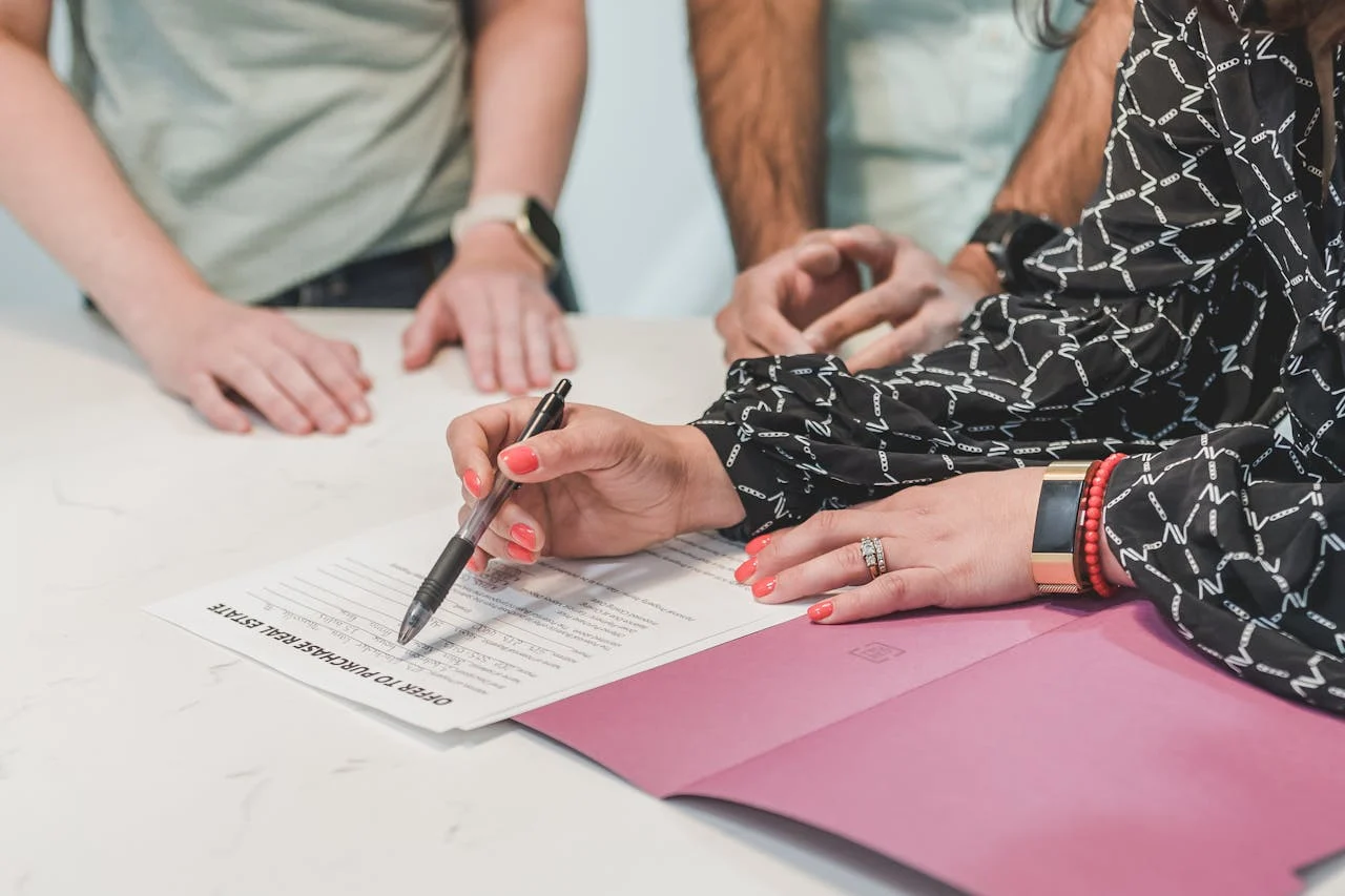 A person is signing a document on a table while three others observe, with a folder and pen visible.