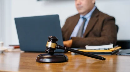 A wooden gavel rests on a desk in the foreground, while a person in a suit works on a laptop in the background.