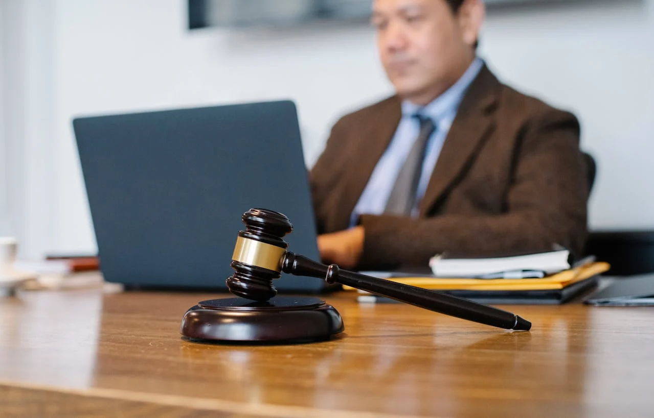 A wooden gavel rests on a desk in the foreground, while a person in a suit works on a laptop in the background.