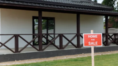 A "Home for Sale" sign is displayed on the lawn in front of a modern single-story house with a porch and dark wood trim.