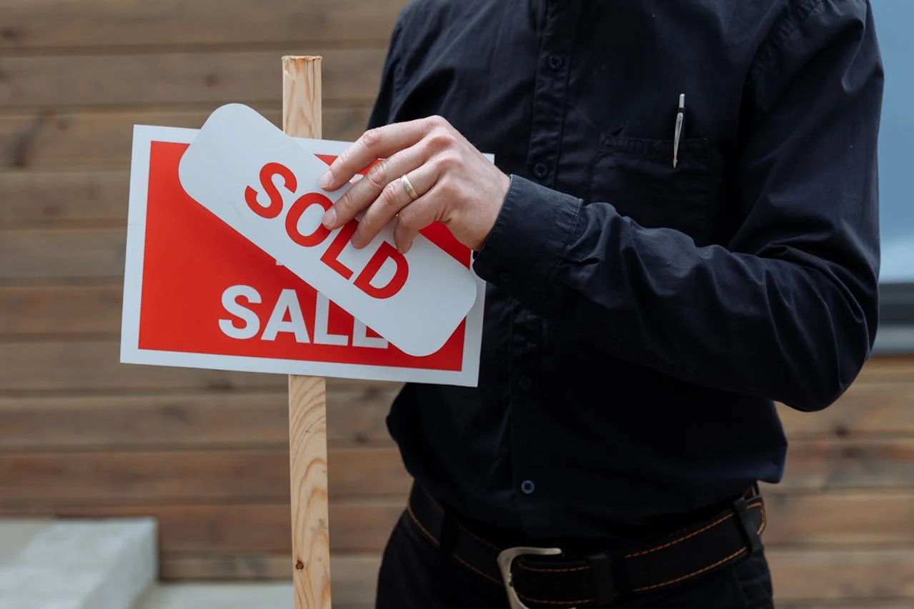 A person places a "SOLD" sign over a "FOR SALE" sign, indicating a sale has been completed.
