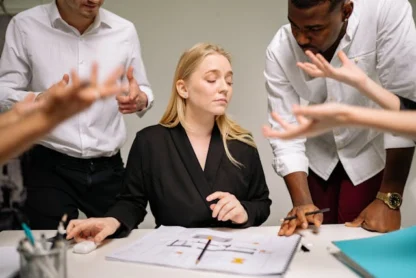 A woman sits at a desk with papers while four colleagues stand around her, gesturing and talking, appearing to have an intense discussion.