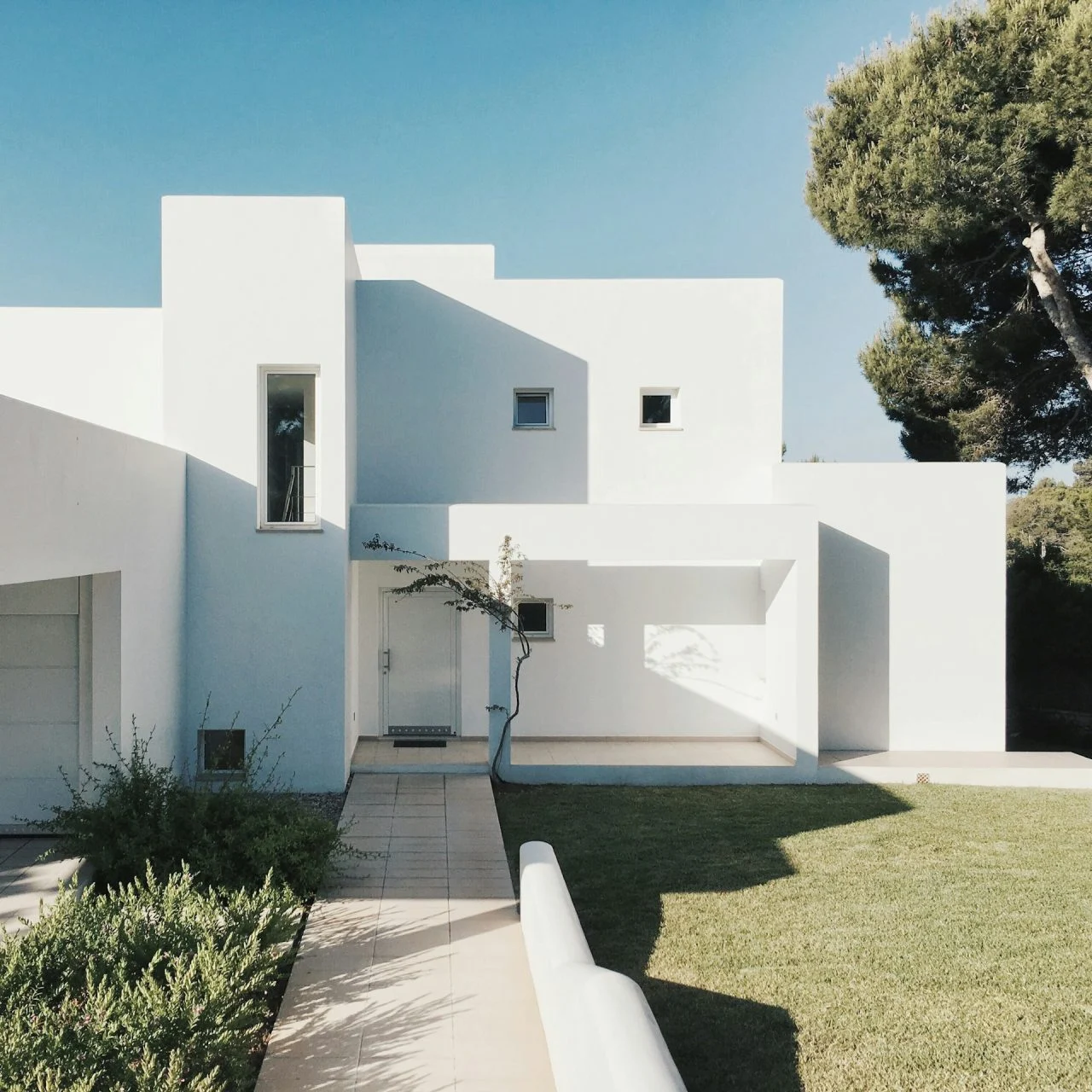 Modern white minimalist house with geometric lines, flat roof, small square windows, and landscaped front yard with grass and shrubbery under a clear blue sky.