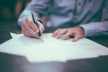 A person in a blue shirt writes on paper with a pen at a desk.