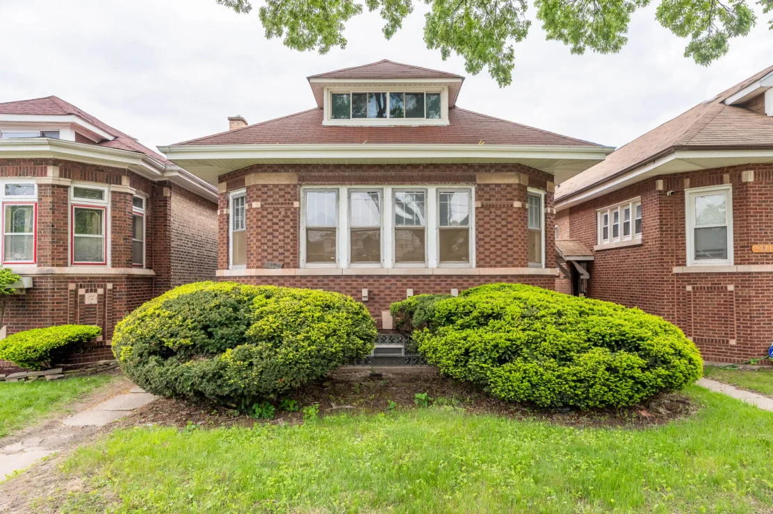 Single-story brick house with large front windows, trimmed bushes, and a small grassy yard, flanked by similar neighboring homes.