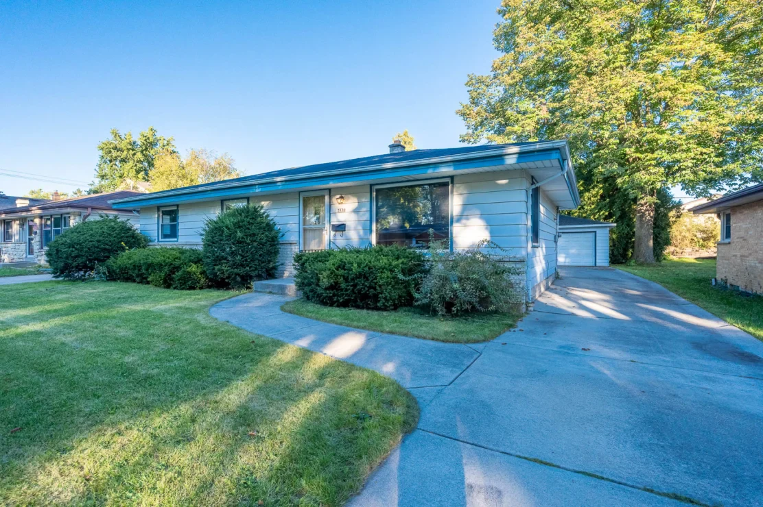Single-story blue house with large windows, surrounded by bushes, a curved driveway, and a detached garage in the background.