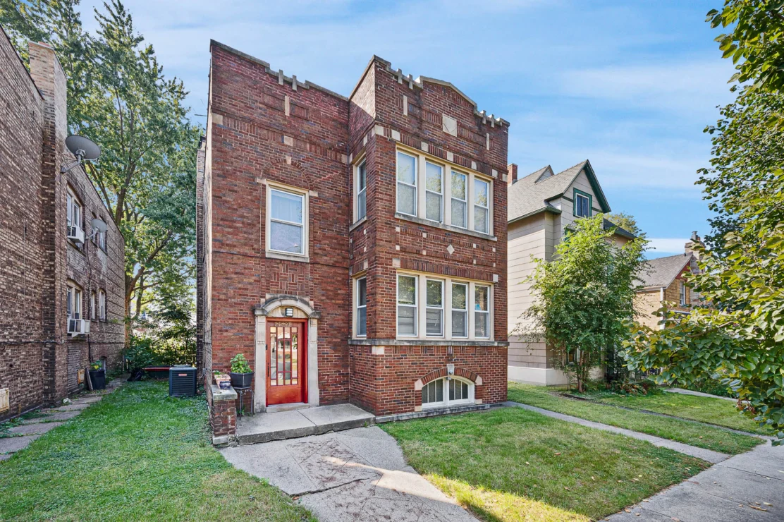 A three-story red brick apartment building with a red door and decorative brickwork, situated between two other houses on a residential street.
