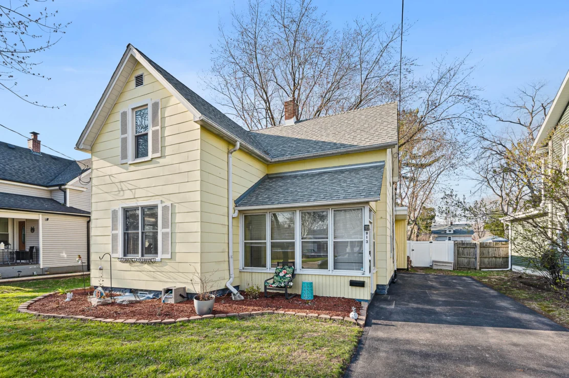 Two-story yellow house with gray roof, enclosed front porch, driveway, manicured lawn, and mulch beds; trees and neighboring houses visible in the background.
