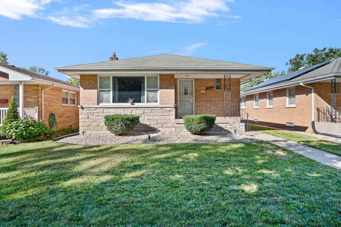 Single-story brick house with a covered front porch, large window, trimmed bushes, stone-lined landscaping, and a well-maintained front lawn.