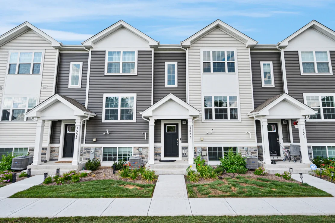 A row of modern townhouses with gray and beige siding, small covered entrances, and landscaped front yards along a sidewalk.