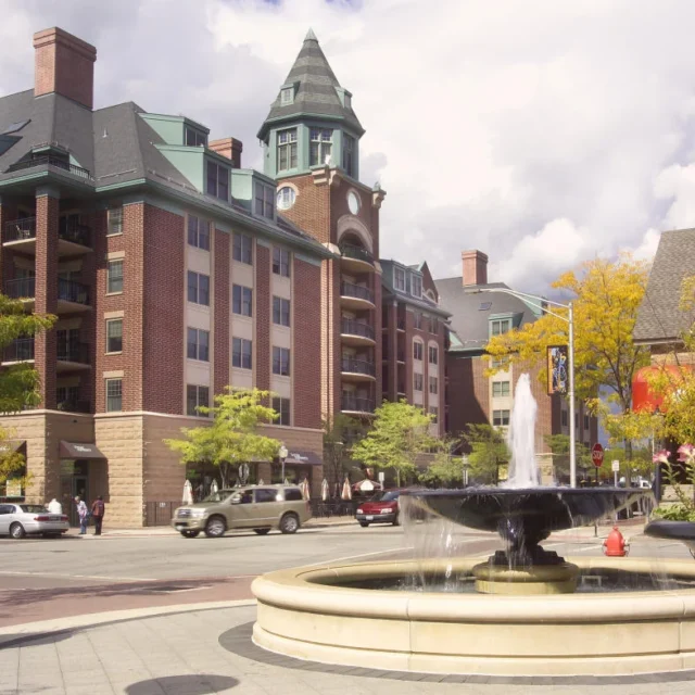 A downtown street scene with brick buildings, a clock tower, parked cars, a round water fountain, and planters with flowers under a partly cloudy sky.