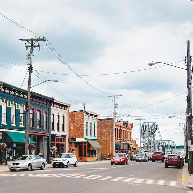 A small town main street with colorful historic buildings, several cars parked and driving, and a bridge visible in the background under a cloudy sky.