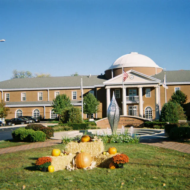 A two-story brick building with columns and a dome, fronted by a sculpture, flag, and autumn-themed decorations with pumpkins and hay bales on a sunny day.