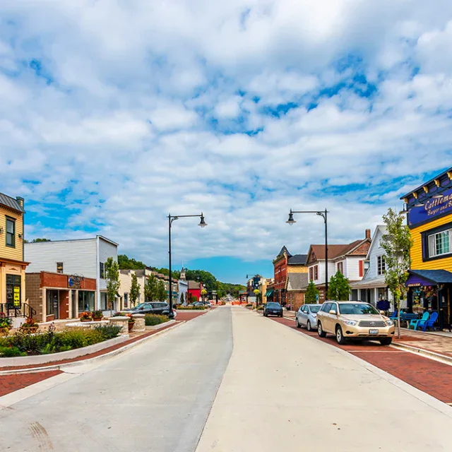 A wide street in a small town with colorful storefronts, parked cars, streetlights, brick sidewalks, trees, and a partly cloudy sky overhead.