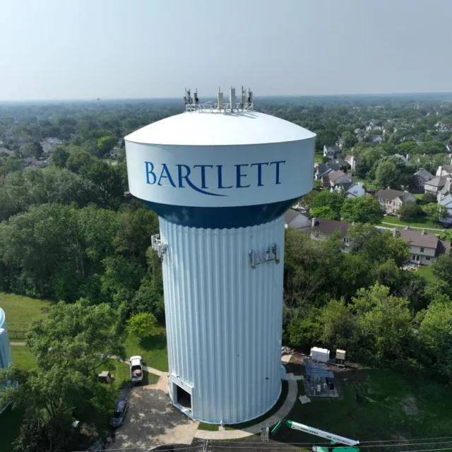 A large white water tower labeled “Bartlett” stands among trees and residential houses on a clear day.