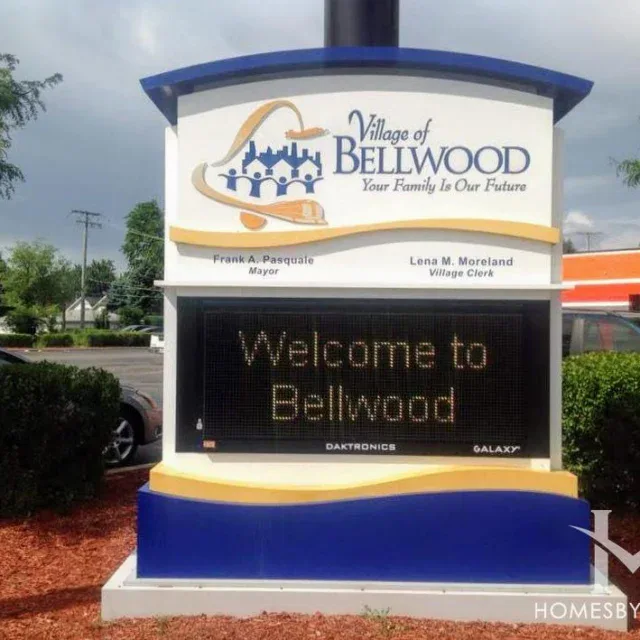 A digital sign reads “Welcome to Bellwood” with the Village of Bellwood logo and mayor and clerk names. The sign is outdoors near a parking lot and landscaping.