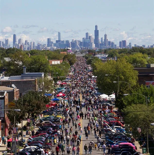 A crowded street fair with parked cars on both sides, people walking down the center, and a city skyline in the background under a partly cloudy sky.