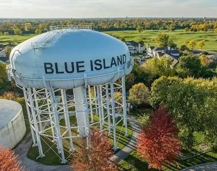 Large water tower labeled “Blue Island” stands surrounded by trees with autumn foliage, residential houses, and a golf course in the background.