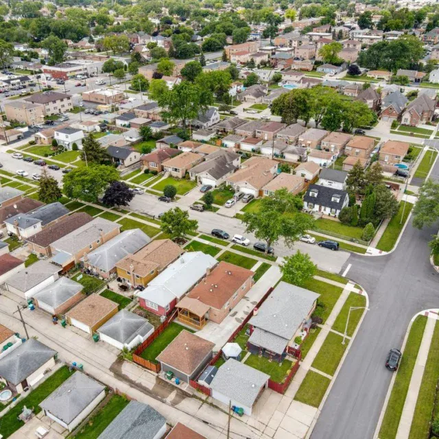 Aerial view of a residential neighborhood with single-family homes, tree-lined streets, sidewalks, and parked cars in an urban area.