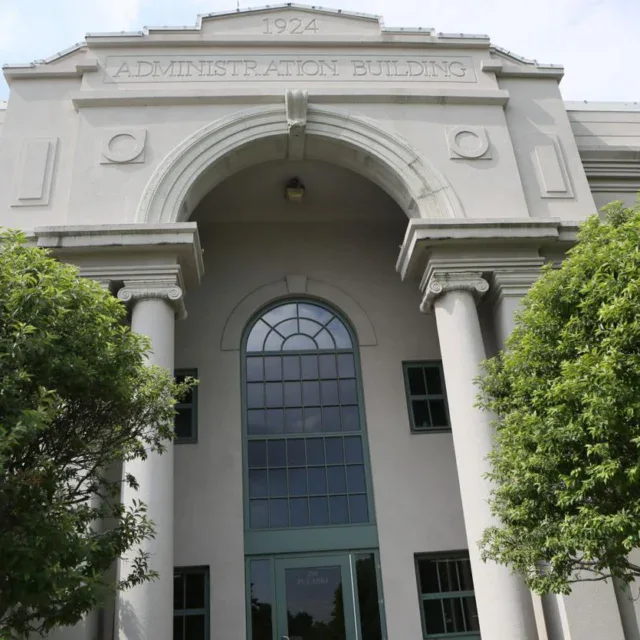 Gray building with columns, labeled “Administration Building 1924” above the entrance, partially obscured by trees.