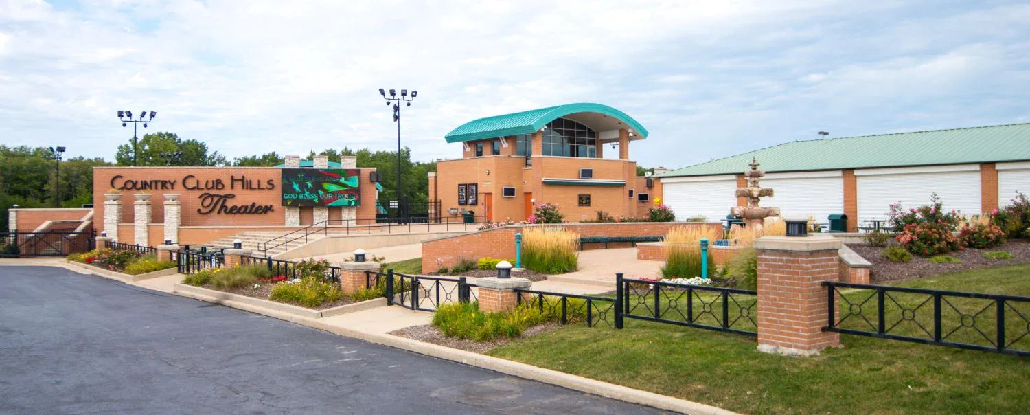 Outdoor Country Club Hills Theater with brick sign, landscaped garden, water fountain, and a modern building with green roof under a partly cloudy sky.
