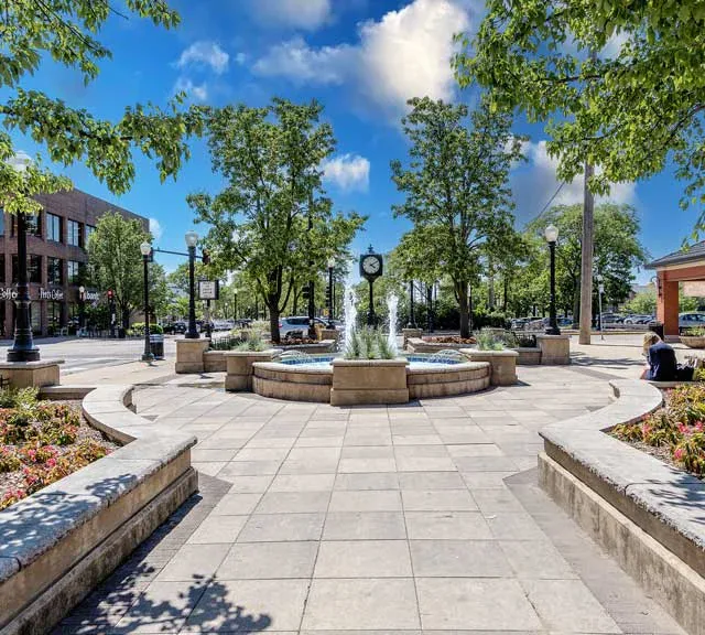 A paved town square with a central fountain, a clock on a pole, benches, flower beds, and trees. A person sits on the right; buildings and cars are visible in the background under a blue sky.