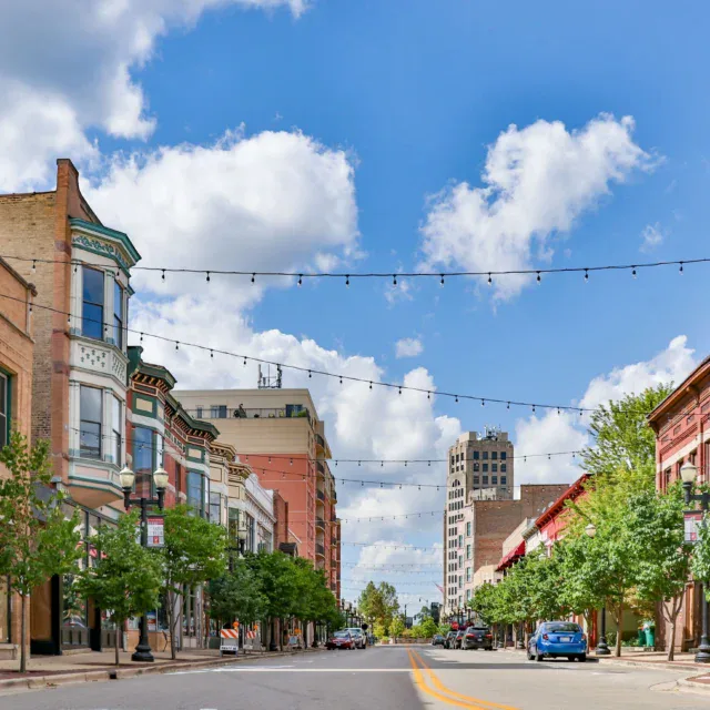 Downtown street view with historic buildings, string lights overhead, parked cars, trees lining sidewalks, and a blue sky with scattered clouds.