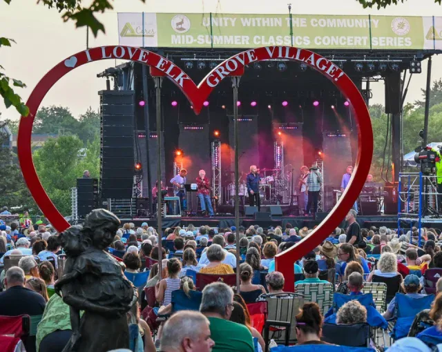 A crowd sits on lawn chairs at an outdoor concert with a band on stage, framed by a large red heart sculpture that reads “I Love Elk Grove Village.”.