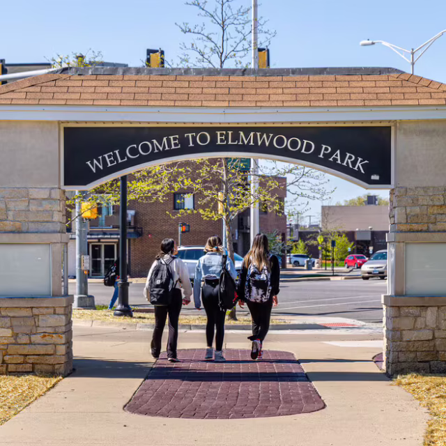 Three people with backpacks walk under a stone and wood archway reading “Welcome to Elmwood Park” on a sunny day.