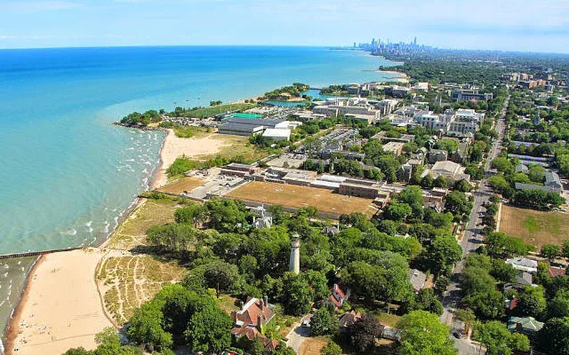 Aerial view of a coastal city with sandy beaches, a lighthouse, residential areas, and a distant skyline under a clear blue sky.