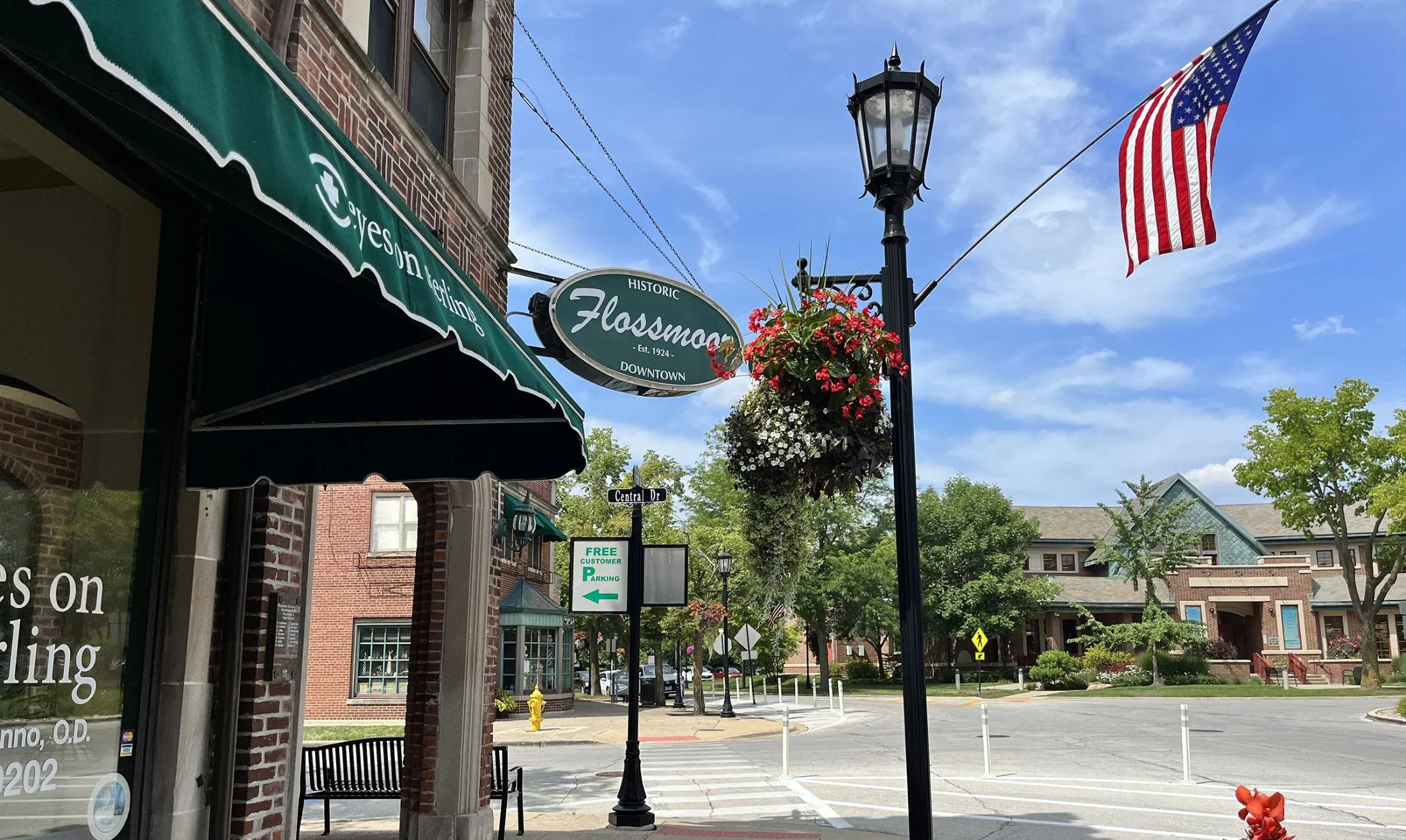 Street view of downtown Flossmoor with a hanging sign, American flag, lamp post, awning, and hanging flowers; buildings and trees line the street on a sunny day.