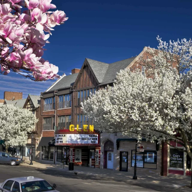 A street lined with blooming pink and white trees, parked cars, and a brick building with a theater marquee that reads “GLEN” under a clear blue sky.