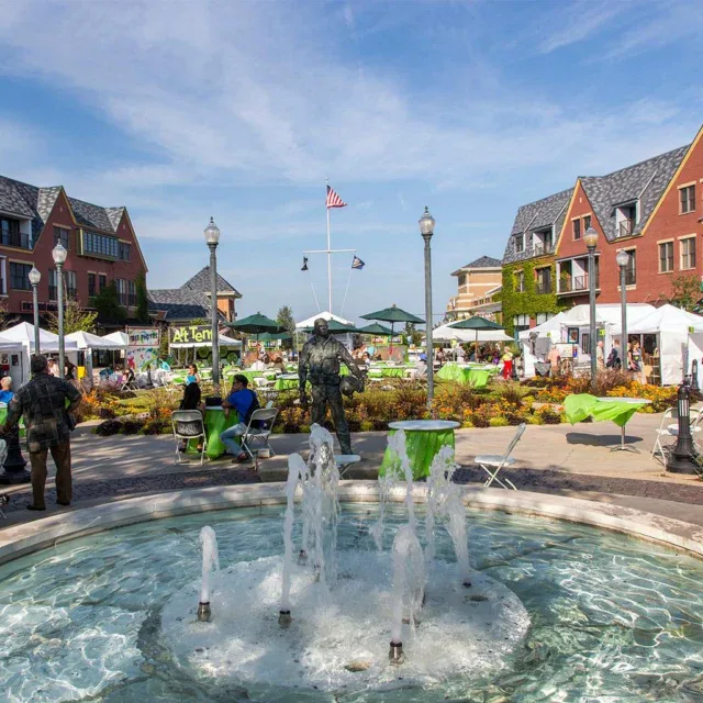 A small fountain in the foreground faces a lively outdoor market with white tents, people sitting at tables, and red brick buildings in the background under a clear blue sky.