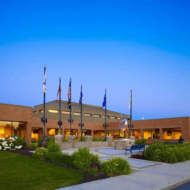 A modern brick building with several flagpoles out front, landscaped greenery, and a clear blue sky at dusk.