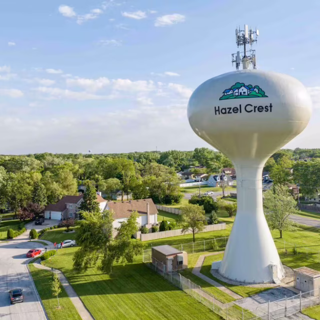 A large white water tower labeled “Hazel Crest” stands on a grassy lot amid houses, trees, and roads under a clear blue sky.