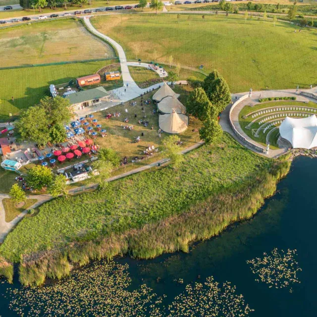 Aerial view of a lakeside park with open grassy areas, outdoor seating with umbrellas, a building, paths, and a round white tent near the water.