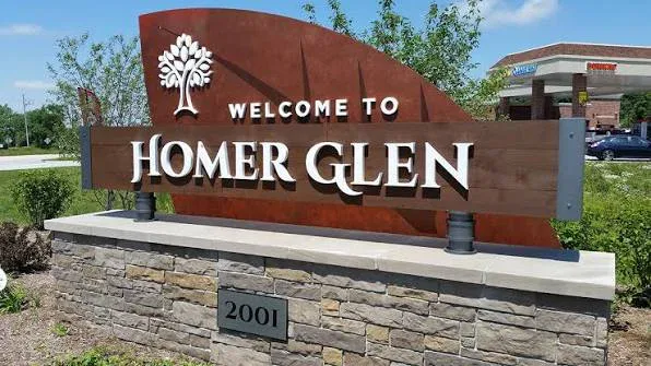 A stone and metal sign reads “Welcome to Homer Glen” with a tree graphic, set near a street and a gas station, with the number 2001 below.