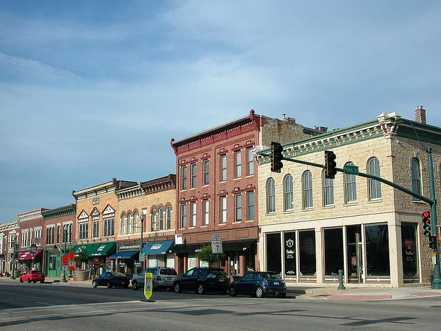 Row of historic brick and stone storefronts along a small town street, with parked cars and a traffic light in the foreground under a clear sky.
