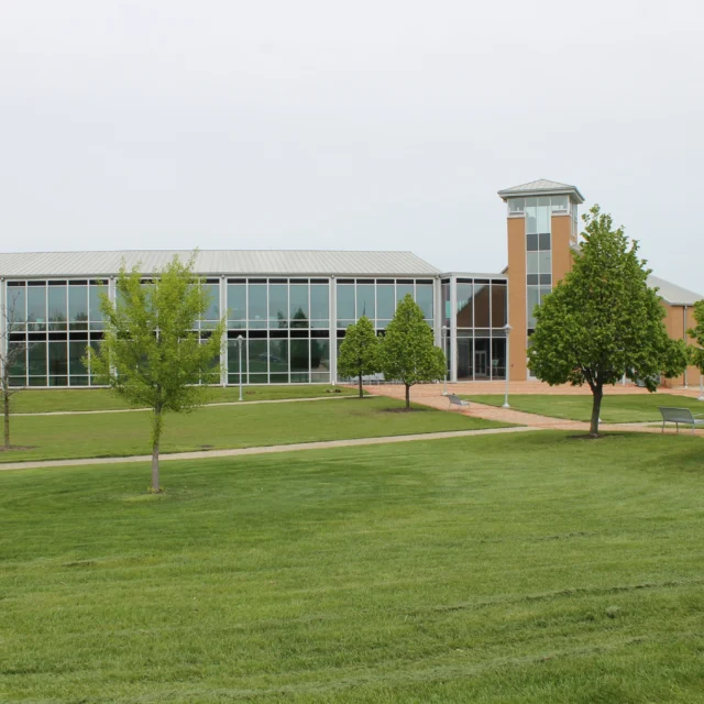 A modern building with large glass windows and a clock tower stands behind a lawn with scattered trees on a cloudy day.