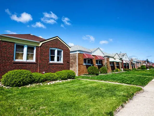 Row of single-story brick houses with well-kept lawns and bushes on a sunny day, viewed from the sidewalk.