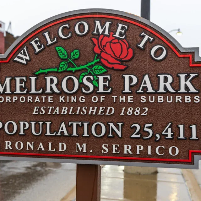 Brown welcome sign for Melrose Park with a red rose, population 25,411, established 1882, and Ronald M. Serpico’s name; street and buildings in the background.