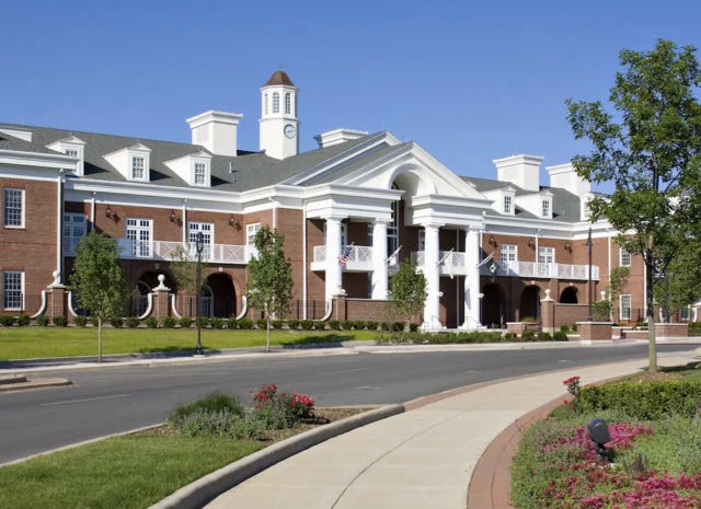 Large red-brick municipal building with white columns and cupola, lined with trees and a curved sidewalk, sign reads “Village of New Lenox.”.