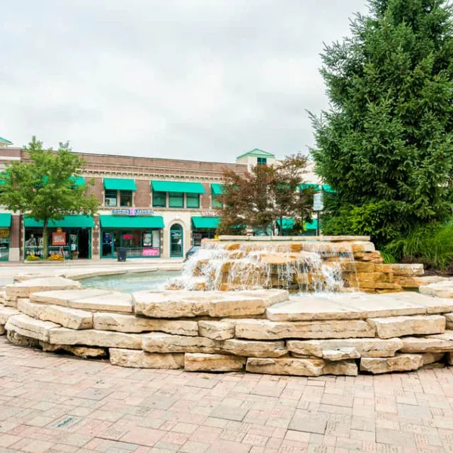 A stone fountain with cascading water sits in a plaza, surrounded by trees and shops with green awnings on a cloudy day.