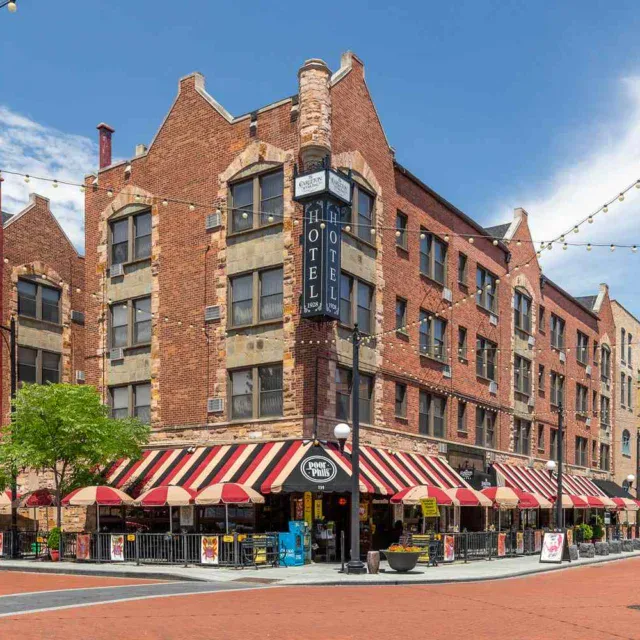 A four-story red brick building with striped awnings houses a hotel and street-level businesses on a sunny day, with string lights crossing the street and outdoor seating visible.