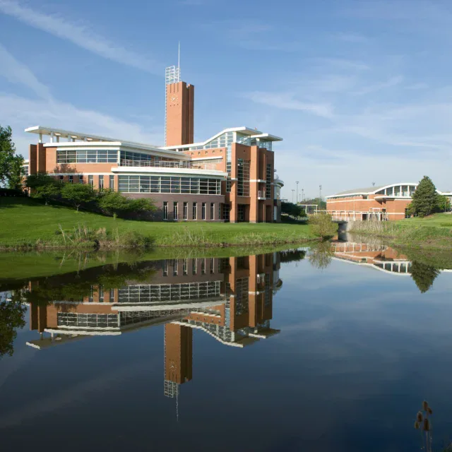 A modern brick building with a tall tower is reflected in a calm pond, surrounded by grass and trees under a blue sky.