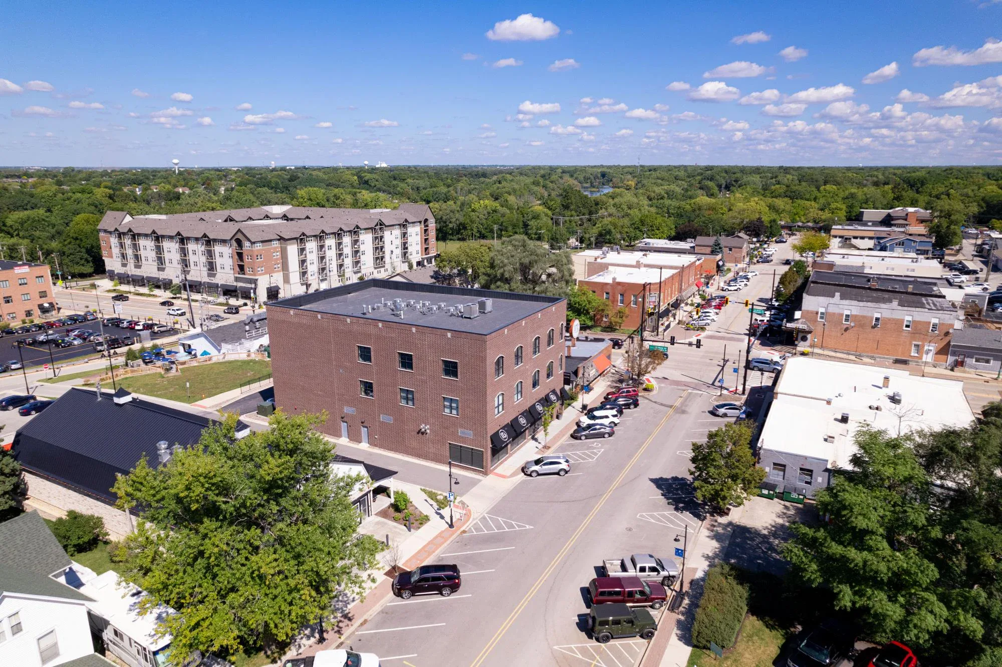 Aerial view of a small town with brick buildings, tree-lined streets, parked cars, and a mix of commercial and residential areas under a clear, sunny sky.