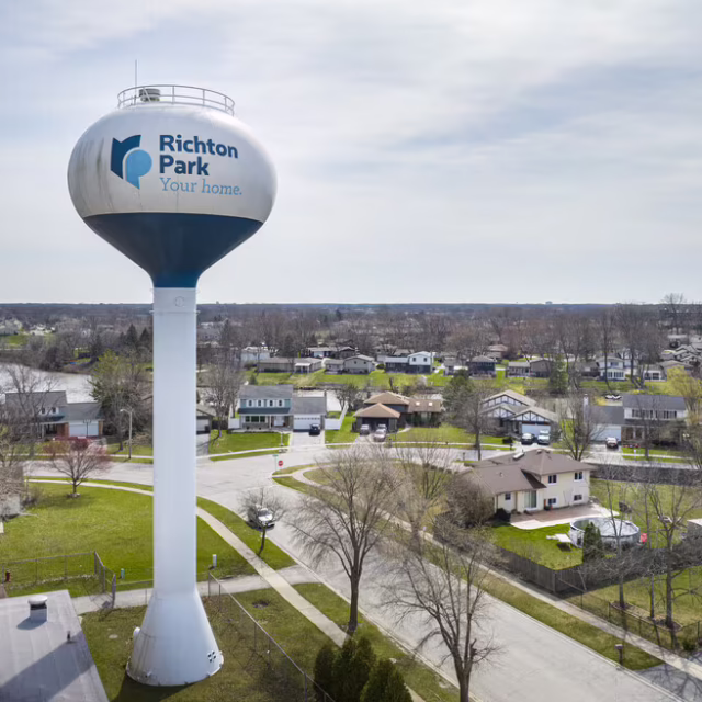 A large water tower labeled “Richton Park Your home” stands over a suburban neighborhood with houses, trees, and roads on a cloudy day.
