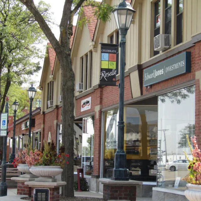 A row of brick storefronts with large windows, business signs, sidewalk planters, benches, and street lamps on a sunny day.